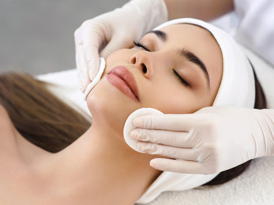 Close up of hands of skillful beautician cleaning and touching female face with cotton pad or sponge. The woman is lying and relaxing. Her eyes are closed with pleasure